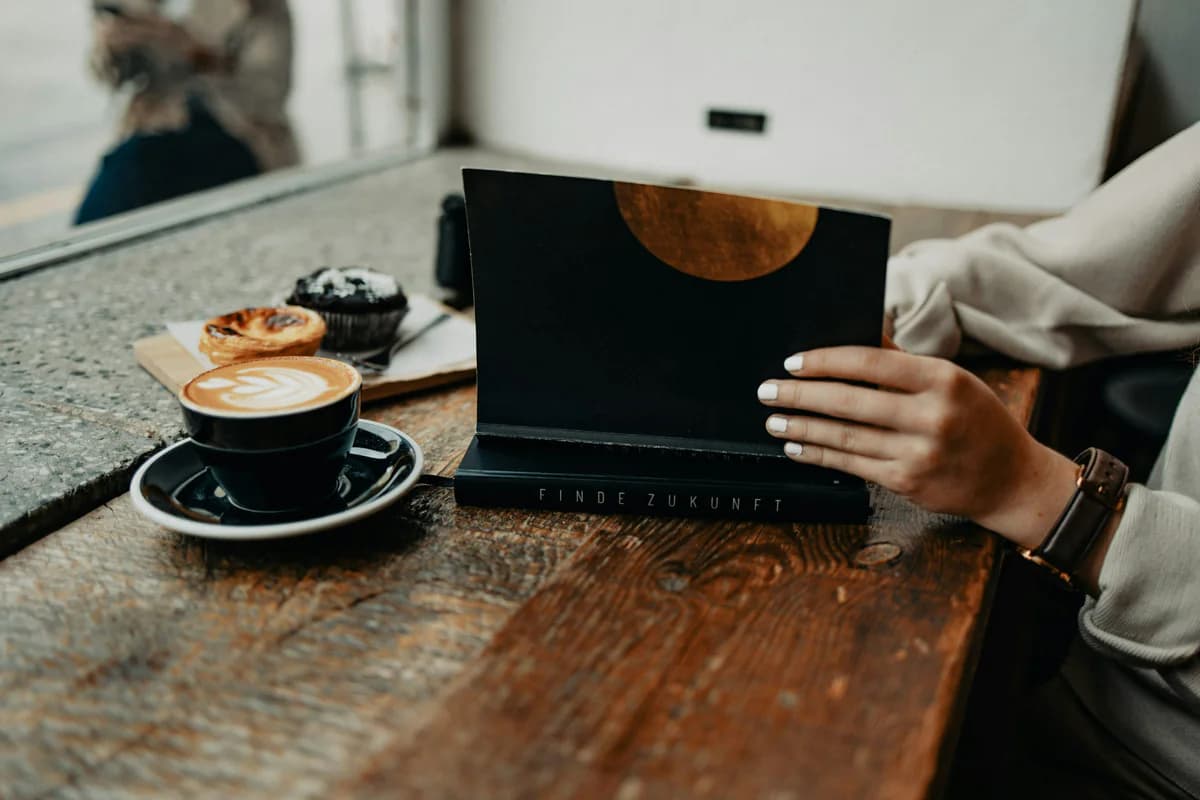 Person holding a book beside a laptop and coffee
