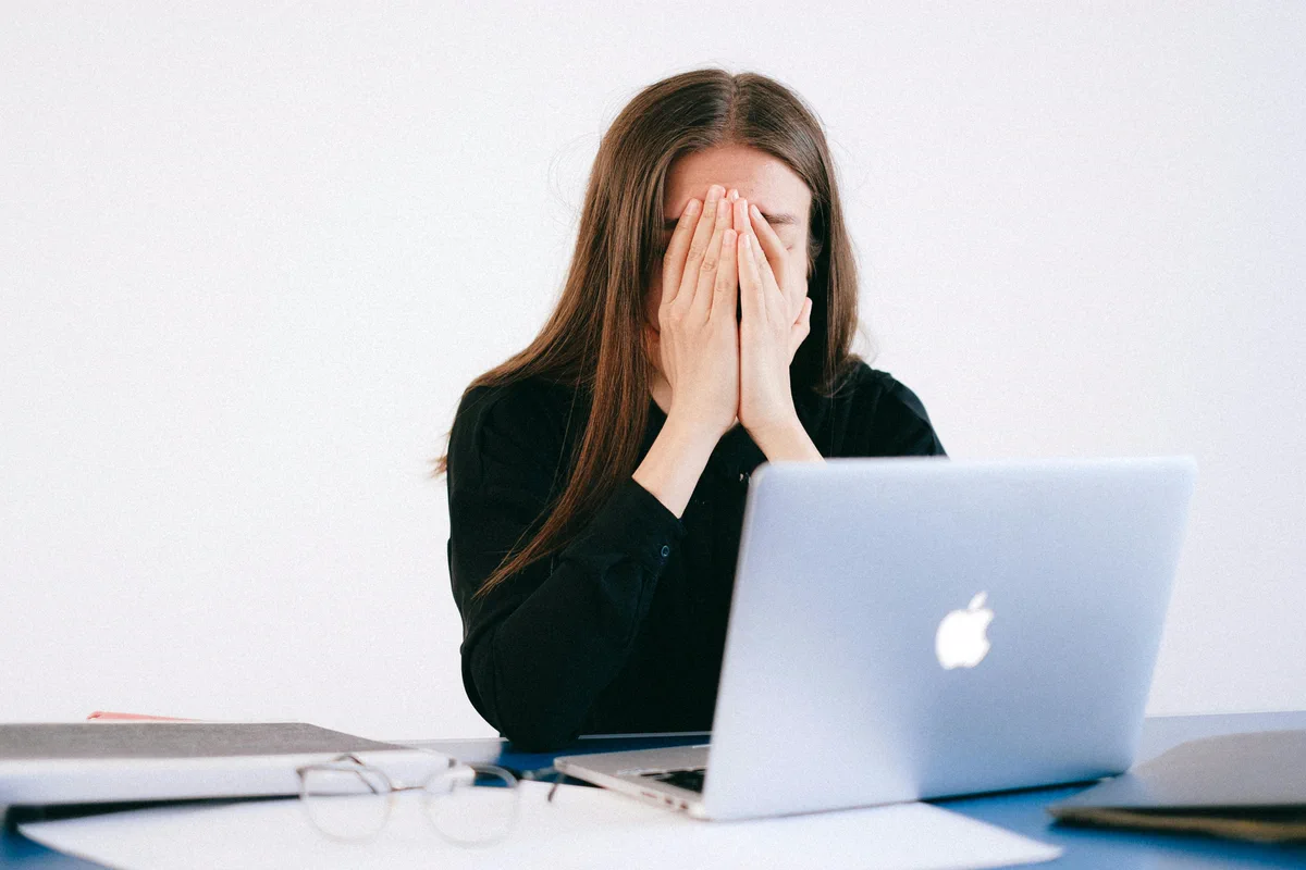A woman covers her face while seated behind a laptop